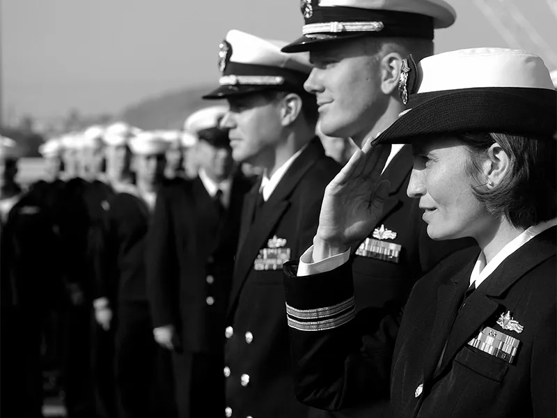 Naval officers in dress whites standing in line, one saluting, in black and white