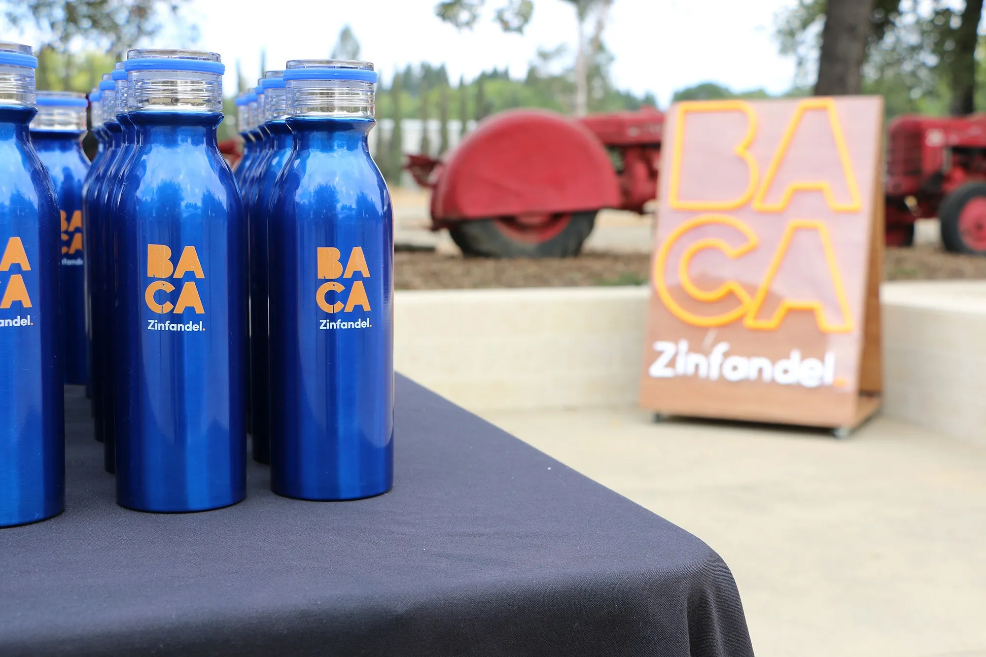 Blue BACA water bottles lined up on a table outdoors, a BACA sign behind them