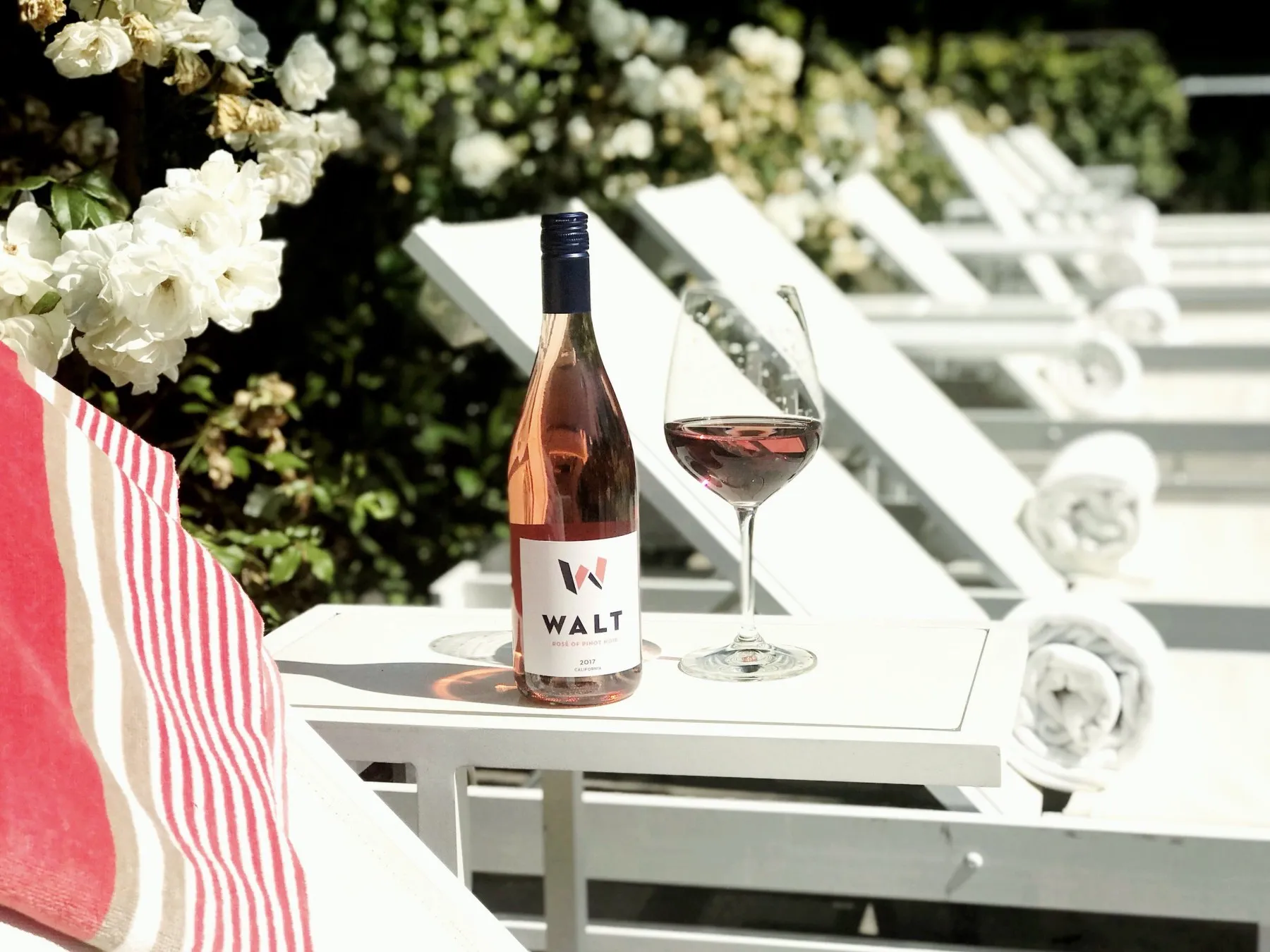 A bottle of WALT rosé and a glass on a table by the pool, white roses behind